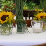 Sommerliche Tischdeko mit Sonnenblumen, Glasvasen und Windlicht auf weißer Tischdecke